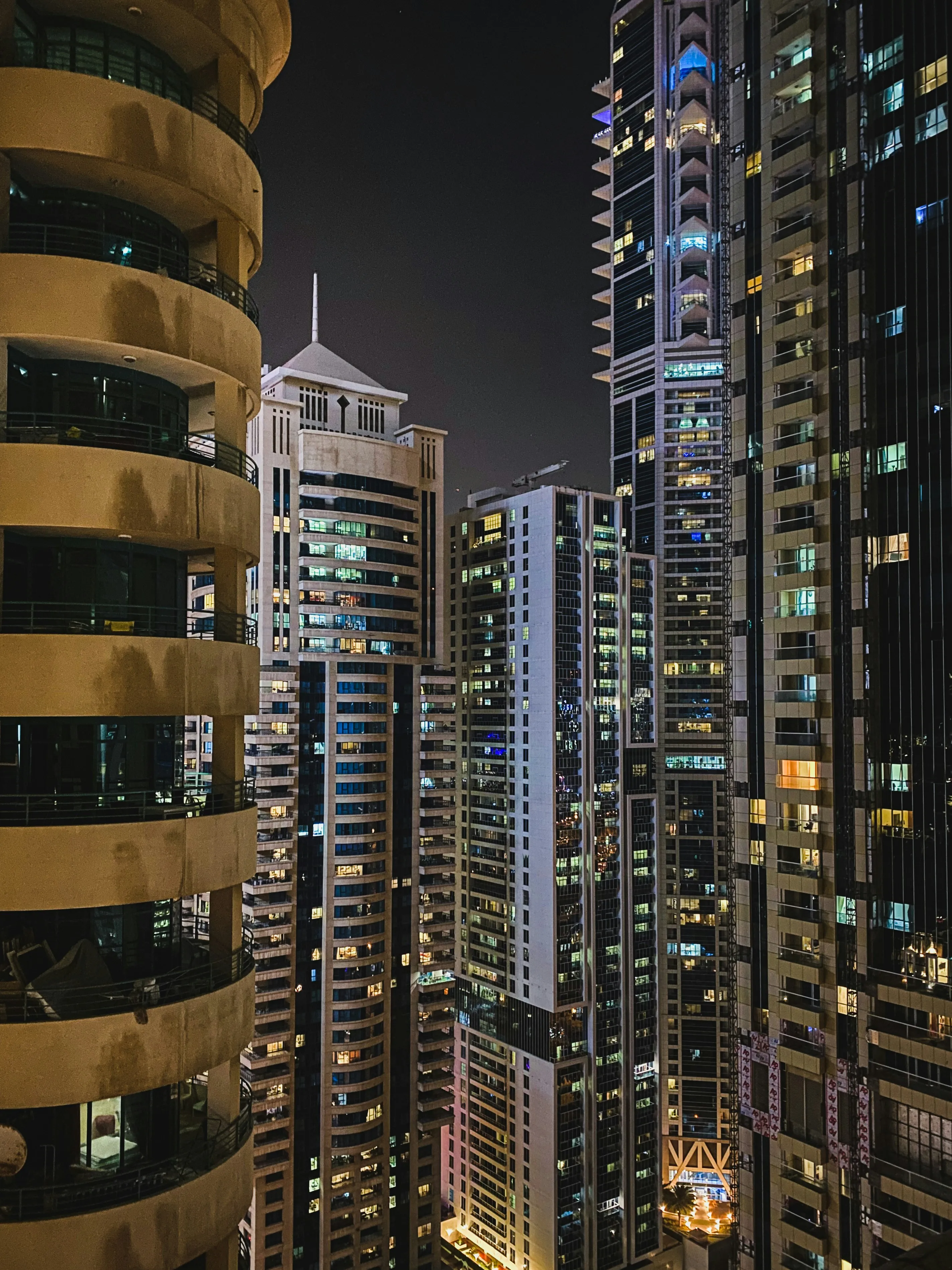 Nocny widok na zwarte wieżowce Dubaju z oświetlonymi oknami i balkonami, miejskie światła i gęsta zabudowa.